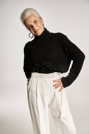 mujer de negocios de mediana edad feliz en elegante traje casual elegante posando con las manos en las caderas en gris