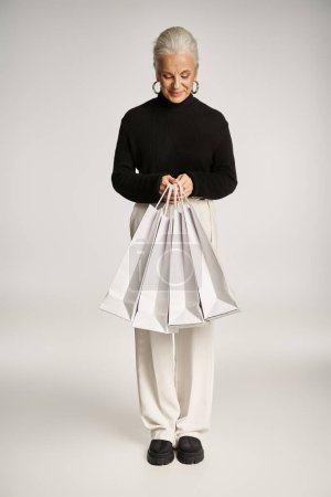 Foto de Alegre mujer de mediana edad en traje elegante y pendientes de aro de pie con bolsas de compras en gris - Imagen libre de derechos