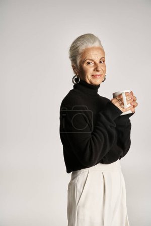 pleased middle aged woman in black turtleneck sweater holding cup of coffee on grey backdrop
