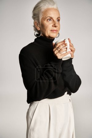 charming middle aged woman in black turtleneck sweater holding cup with coffee on grey backdrop
