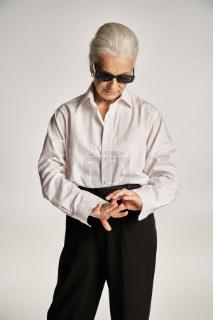 stylish middle aged woman in white shirt, black pants and sunglasses looking at rings on fingers