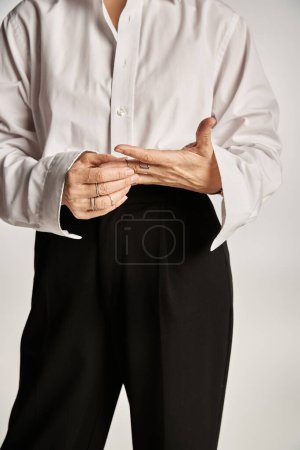 cropped middle aged woman in white shirt, black pants touching rings on fingers on grey backdrop