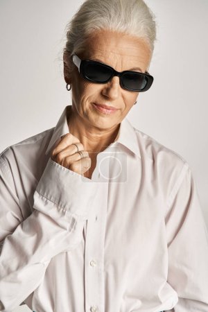 modische Frau mittleren Alters in edlem weißen Hemd und Sonnenbrille posiert auf grauem Hintergrund