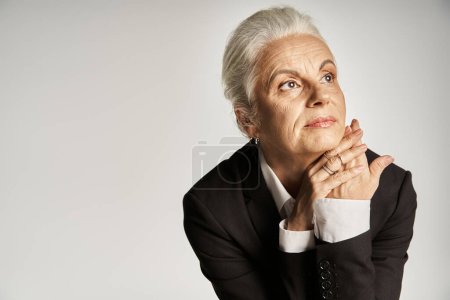 hermosa y gris mujer de mediana edad en traje elegante posando sobre fondo gris