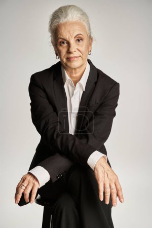 schöne und grauhaarige Frau mittleren Alters in eleganter Kleidung posiert auf grauem Hintergrund