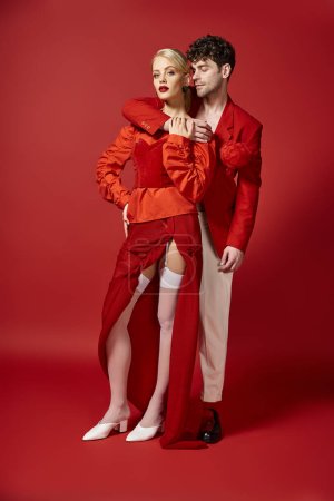 full length of handsome man embracing woman in elegant attire on red background, stylish couple