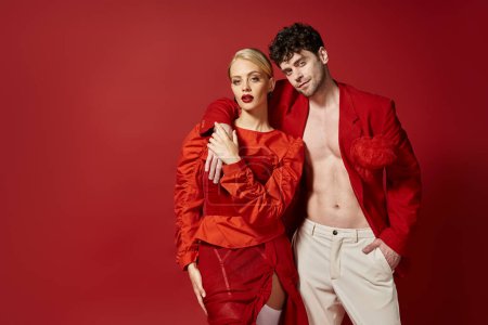 stylischer Mann in rotem Blazer und weißer Hose posiert, während er der blonden Frau die Hand um den Hals legt