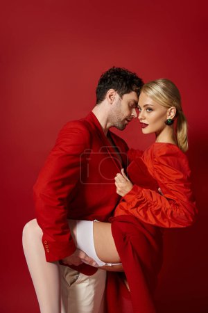 schöner Mann hebt Bein der verführerischen blonden Frau in sexy weißen Strümpfen auf rotem Hintergrund