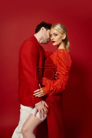 Téléchargez les photos : Bel homme touchant jambe de séduisante femme blonde en bas sexy sur fond rouge, de la mode - en image libre de droit