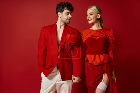 alegre rubia mujer cogida de la mano del hombre guapo en elegante atuendo sobre fondo rojo, moda
