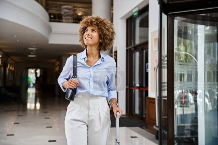Téléchargez les photos : Joyeuse voyageuse afro-américaine marchant à l'entrée du hall de l'hôtel avec sa valise - en image libre de droit