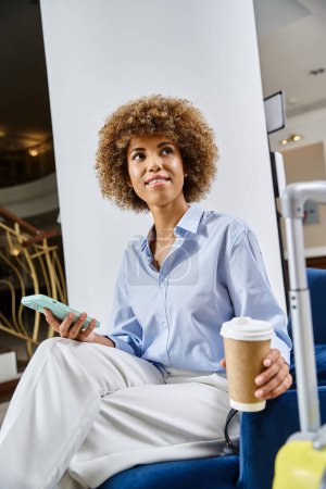 Mujer afroamericana relajada y soñadora con café para llevar y teléfono inteligente esperando en el vestíbulo del hotel