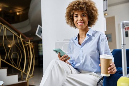 lockige und glückliche Afroamerikanerin mit Coffee to go und Smartphone, die in der Hotellobby wartet