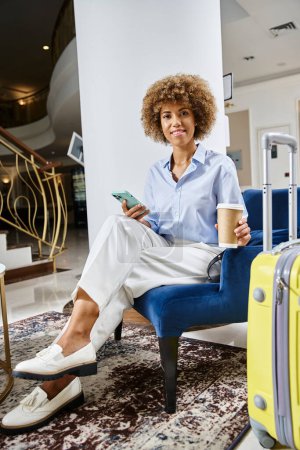Téléchargez les photos : Femme afro-américaine détendue et souriante avec café à emporter et smartphone dans le hall de l'hôtel - en image libre de droit