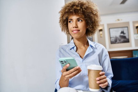 Téléchargez les photos : Heureuse femme afro-américaine bouclée avec café à emporter et smartphone assis dans le hall de l'hôtel - en image libre de droit