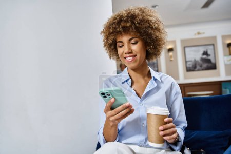 happy curly african american woman with coffee to go and using smartphone in hotel lobby