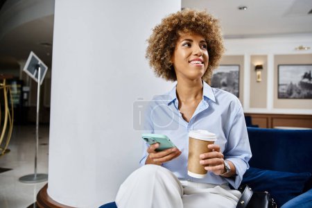 Téléchargez les photos : Heureuse femme afro-américaine bouclée avec du café à emporter et en utilisant un smartphone dans le hall de l'hôtel - en image libre de droit