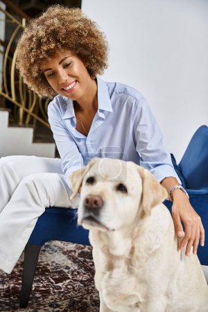 happy african american woman with curly hair sitting and cuddling labrador in pet-friendly hotel