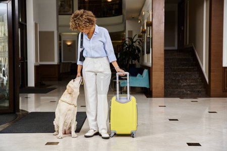 Téléchargez les photos : Voyageuse heureuse avec son Labrador dans une entrée d'hôtel acceptant les animaux domestiques, femme afro-américaine et chien - en image libre de droit