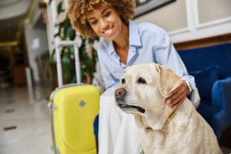 glücklicher Reisender wartet auf Check-in mit Labrador im haustierfreundlichen Hotel, schwarze Frau mit Hund