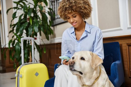 Téléchargez les photos : Touriste heureux avec chien et téléphone dans un hôtel acceptant les animaux, labrador et femme afro-américaine - en image libre de droit