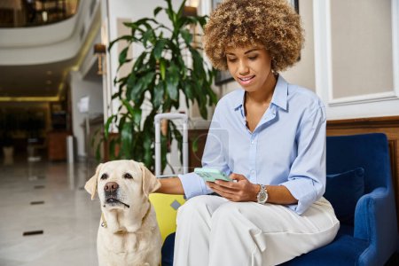Téléchargez les photos : Touriste avec chien et téléphone dans un hôtel acceptant les animaux, labrador et heureuse femme afro-américaine - en image libre de droit