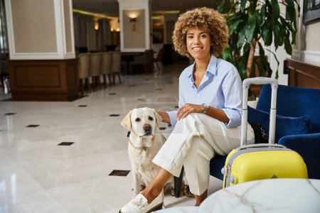 Téléchargez les photos : Heureuse invitée en attente d'enregistrement avec son Labrador dans un hôtel acceptant les animaux de compagnie, femme noire câlin chien - en image libre de droit