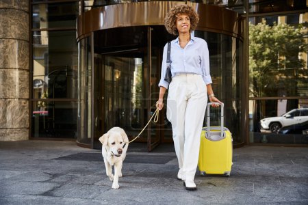 Lockige Afroamerikanerin mit Hund und Gepäck spaziert aus haustierfreundlichem Hotel
