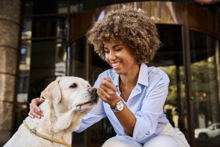 Téléchargez les photos : Femme afro-américaine souriante et frisée câlinant labrador devant un hôtel acceptant les animaux de compagnie - en image libre de droit