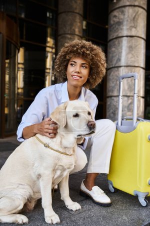 Téléchargez les photos : Femme afro-américaine souriante câlinant labrador près des bagages devant un hôtel acceptant les animaux de compagnie - en image libre de droit