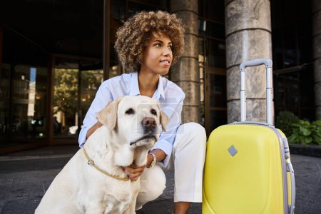 Téléchargez les photos : Joyeuse femme afro-américaine câlinant labrador près des bagages devant un hôtel acceptant les animaux de compagnie - en image libre de droit