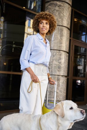 pleased african american woman with dog and luggage standing near entrance of pet friendly hotel