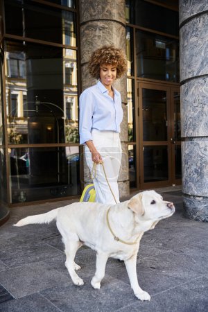 mujer afroamericana complacida con labrador de pie cerca de la entrada del hotel que acepta mascotas