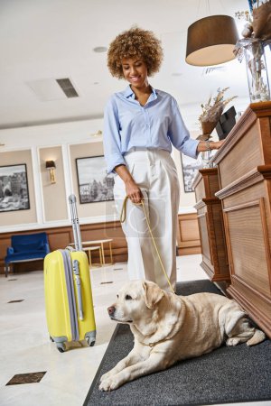 fröhliche afrikanisch-amerikanische Frau mit ihrem Labrador beim Einchecken in einem haustierfreundlichen Hotel an der Rezeption
