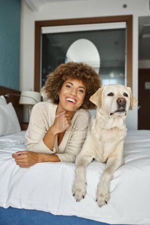 fröhliche afrikanisch-amerikanische Frau mit ihrem Labrador auf einem Bett in einem haustierfreundlichen Hotel liegend, Reisen