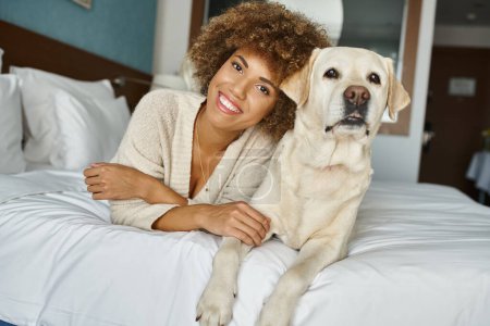 Gleful afrikanisch-amerikanische Frau mit ihrem Labrador liegend auf einem Bett in einem haustierfreundlichen Hotel, Reisen