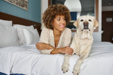 freudige afrikanisch-amerikanische Frau mit ihrem Labrador auf einem Bett in einem haustierfreundlichen Hotel liegend, Reisen