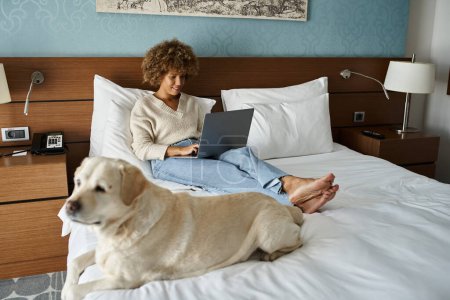 young african american woman working on laptop with her labrador on bed in a hotel room