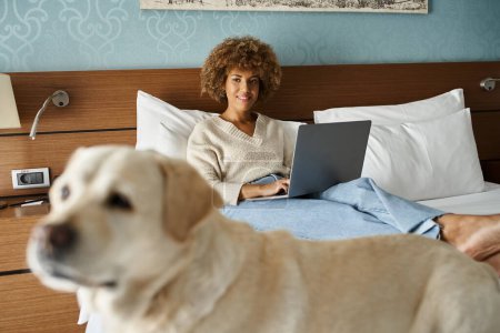 young african american woman working on laptop and looking at labrador on bed in a hotel room