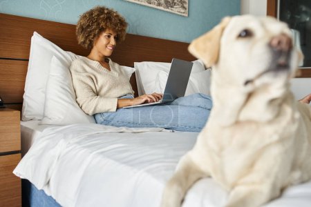 happy young african american woman working on laptop with her labrador on bed in a hotel room