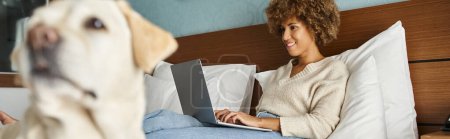 jeune femme afro-américaine travaillant sur ordinateur portable avec son labrador sur le lit dans une chambre d'hôtel, bannière