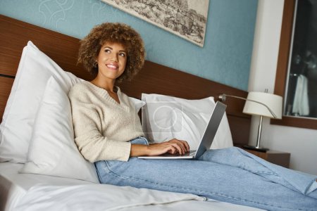 happy and curly african american woman looking away while working on laptop on bed in a hotel room