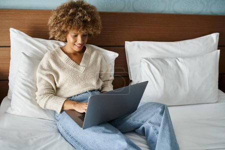 heureuse et frisée afro-américaine souriante tout en travaillant sur un ordinateur portable sur le lit dans une chambre d'hôtel