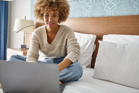 positive young african american woman with curly hair working on laptop on bed in hotel room