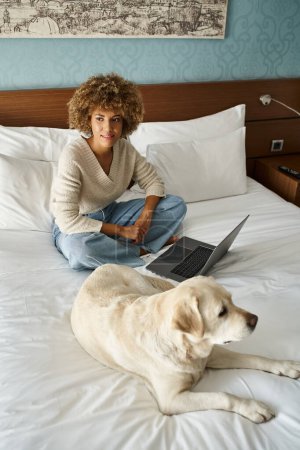 soñadora joven afroamericana sentada en la cama cerca de labrador y laptop en habitación de hotel moderna