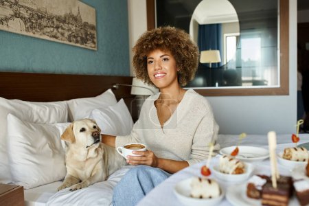 Téléchargez les photos : Joyeuse femme afro-américaine tenant cappuccino et câlinant son chien labrador dans un hôtel acceptant les animaux de compagnie - en image libre de droit