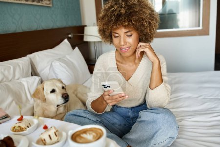heureuse femme afro-américaine tenant cappuccino et utilisant un smartphone près de labrador dans la chambre d'hôtel