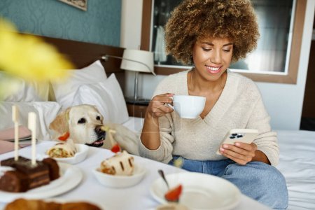 heureuse femme afro-américaine tenant cappuccino et utilisant un smartphone près de labrador dans la chambre d'hôtel