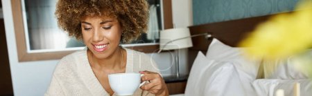 heureuse jeune femme afro-américaine avec les cheveux bouclés savourant cappuccino dans la chambre d'hôtel, bannière