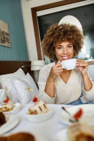 mujer afroamericana feliz sosteniendo taza y sentado en la cama en la habitación de hotel, servicio de habitaciones y comodidad